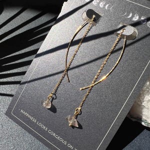 May include: A pair of gold earrings with a delicate chain and a clear crystal stone hanging from each. The earrings are displayed on a black card with the text "Happiness looks gorgeous on you."