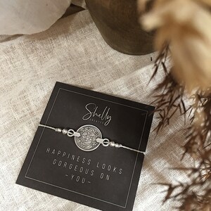 Puede incluir: Un brazalete de plata con un dije redondo que dice "Happiness looks gorgeous on you". El brazalete est&aacute; sobre una tarjeta negra con el texto "Shelly Creates" en blanco.