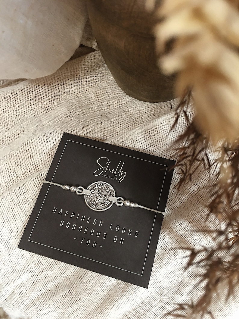 Puede incluir: Un brazalete de plata con un dije redondo que dice "Happiness looks gorgeous on you". El brazalete est&aacute; sobre una tarjeta negra con el texto "Shelly Creates" en blanco.