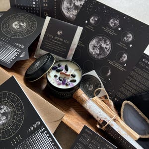 Könnte beinhalten: Eine Sammlung von schwarz-weißen Artikeln zum Thema Himmelskörper, darunter ein Januar-Kalender, eine Full Moon Tee-Packung, eine Kerze mit violetten und schwarzen Steinen, ein Glasröhrchen mit Salzen und einem Holzstab sowie Mondphasenkarten.