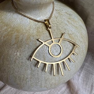 May include: A gold necklace with a large eye-shaped pendant. The pendant is made of metal and has a detailed design.