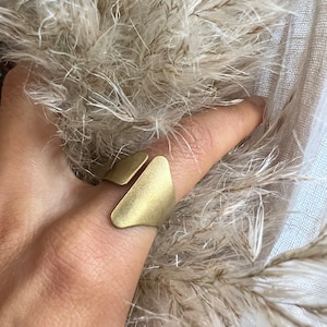 Könnte beinhalten: Ein goldfarbener Ring mit geometrischem Design. Der Ring besteht aus zwei dreieckigen Teilen, die oben miteinander verbunden sind. Der Ring wird an einem Finger getragen.