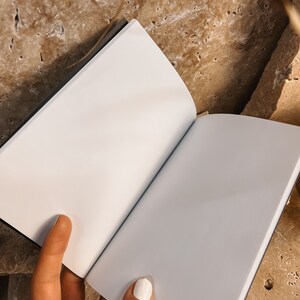 Puede incluir: Un cuaderno blanco en blanco con una cubierta gris oscuro est&aacute; abierto y sobre una superficie texturizada de color marr&oacute;n claro. Una mano sostiene el cuaderno abierto.