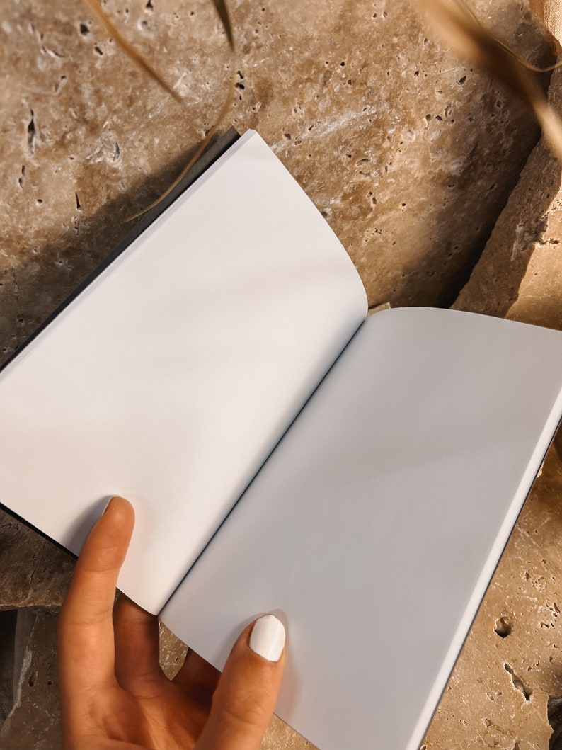 Puede incluir: Un cuaderno blanco en blanco con una cubierta gris oscuro est&aacute; abierto y sobre una superficie texturizada de color marr&oacute;n claro. Una mano sostiene el cuaderno abierto.