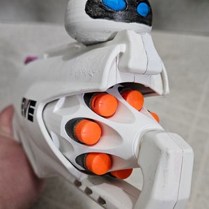 May include: A white toy gun with orange and black details, featuring a spherical head with blue eyes and black accents. The word "EVE" is printed on the side. The toy is a replica of the character Eve from the movie Wall-E.