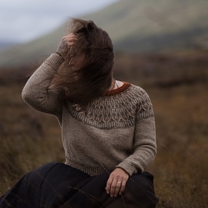 May include: A person wearing a brown and beige jumper with a brown and orange patterned design sits in a field of long grass. The person's hair is blowing in the wind.