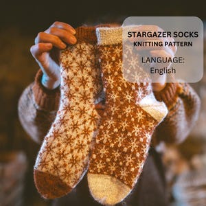 Könnte beinhalten: Ein Paar gestrickte Socken mit einem Schneeflockenmuster in Braun- und Cremetönen. Die Socken werden hochgehalten, um das Design zu präsentieren. Der Text auf dem Bild lautet "STARGAZER SOCKS KNITTING PATTERN LANGUAGE: English."