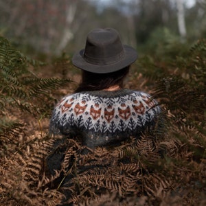 May include: A person wearing a brown hat and a green sweater with a fox pattern sits in a forest setting. The sweater has a white and brown pattern with fox faces.