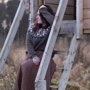May include: A woman wearing a brown sweater with a white and brown patterned design sits on a wooden ladder. She is wearing a brown and black plaid skirt. The ladder is leaning against a wooden structure.