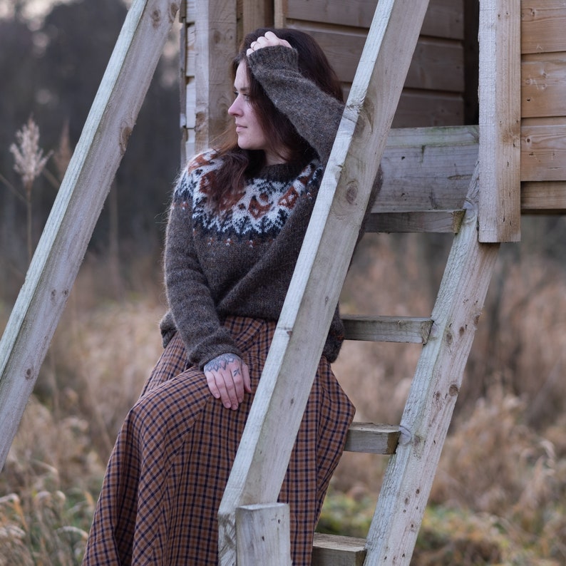 May include: A woman wearing a brown sweater with a white and brown patterned design sits on a wooden ladder. She is wearing a brown and black plaid skirt. The ladder is leaning against a wooden structure.