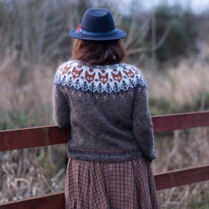 May include: A woman wearing a brown sweater with a fox pattern and a black hat. She is standing in front of a wooden fence.