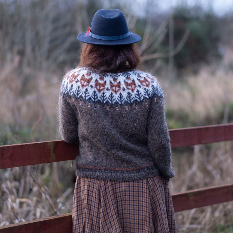 May include: A woman wearing a brown sweater with a fox pattern and a black hat. She is standing in front of a wooden fence.