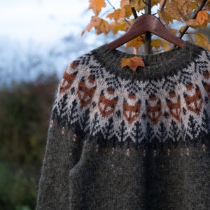 May include: A green, brown, white, and orange knitted sweater with a traditional Scandinavian pattern. The sweater is hanging on a wooden hanger.