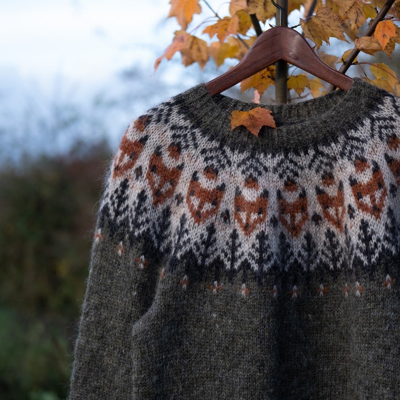 May include: A green, brown, white, and orange knitted sweater with a traditional Scandinavian pattern. The sweater is hanging on a wooden hanger.