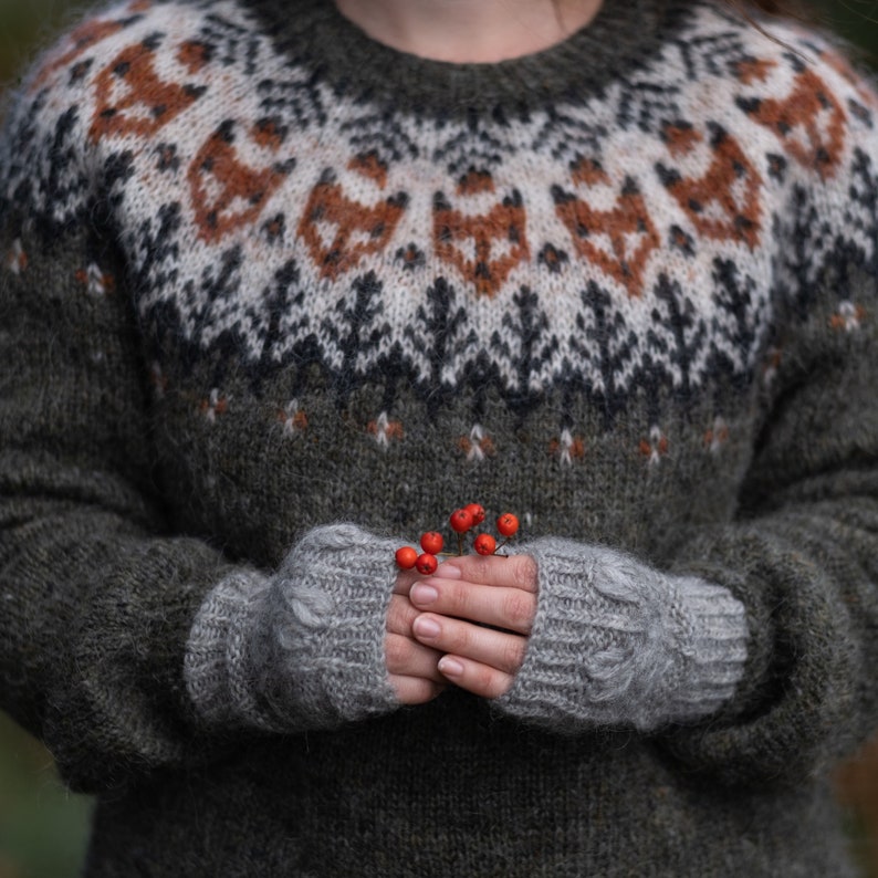 May include: A close-up of a person wearing a gray and brown knitted sweater with a traditional Scandinavian pattern. The person is holding a sprig of red berries in their hands, which are also wearing gray knitted fingerless gloves.