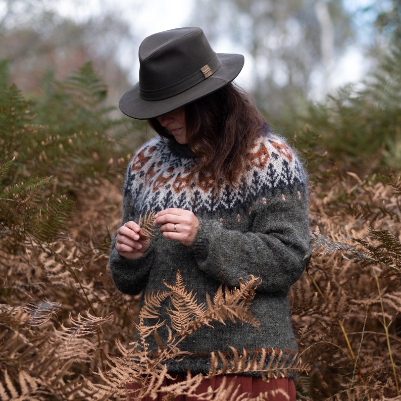 May include: A woman wearing a green sweater with a brown and white patterned design, a brown felt hat, and a brown skirt. She is standing in a field of brown ferns.