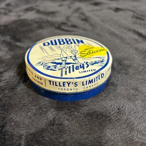 May include: A blue and white round tin with the text "DUBBIN" in yellow letters. The tin has a picture of a person wearing a coat and boots walking in the rain. The text "WITH Silicone" is in a yellow circle on the tin. The text "Tilley's LIMITED" is in blue letters on the tin. The text "TILLEY'S LIMITED" is in blue letters on the tin. The text "TORONTO, CANADA" is in blue letters on the tin.