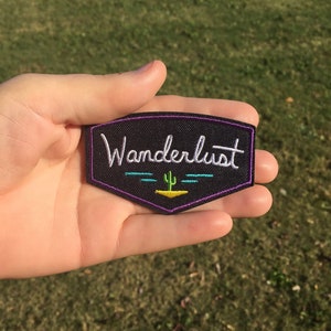 May include: A black embroidered patch with the word "Wanderlust" in white script. The patch features a purple border and a desert scene with a cactus and sun design in teal and yellow.