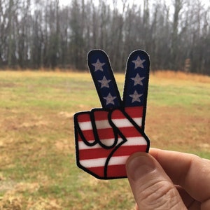 May include: A peace sign patch with the American flag design. The patch is red, white, and blue with white stars on a blue background.