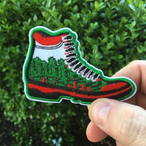 May include: Green and red embroidered patch of a hiking boot with white laces.