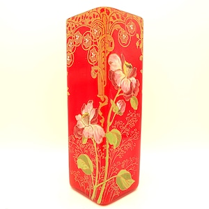 May include: A rectangular red glass vase with a floral design. The vase features a gold filigree pattern at the top and a painted floral motif with pink and white flowers and green leaves. The vase is approximately 10 inches tall.