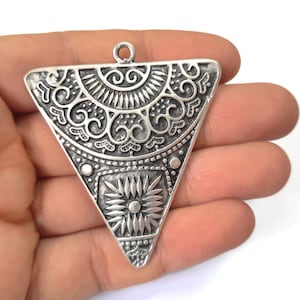 Puede incluir: Colgante triangular de plata con un diseño intrincado y ornamentado. El colgante presenta un patrón circular con motivos florales y un estallido de estrellas central.