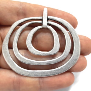 May include: A silver-toned pendant with a layered, open-circle design. The pendant features three nested, irregular oval shapes, with a small loop at the top for hanging. The metal has a slightly textured, matte finish.