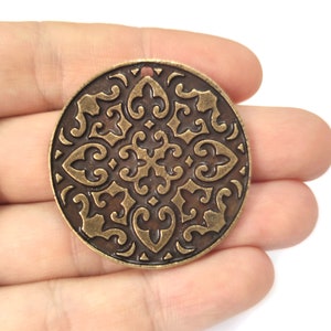 以下が含まれることがあります： 複雑で装飾的なデザインが施された、丸いブロンズ色の金属製のペンダント。ペンダントの上部に吊り下げ用の穴があります。