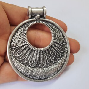 May include: Silver pendant with an intricate, circular design featuring a large open circle in the center. The pendant has a textured surface and a loop at the top for attaching a chain.
