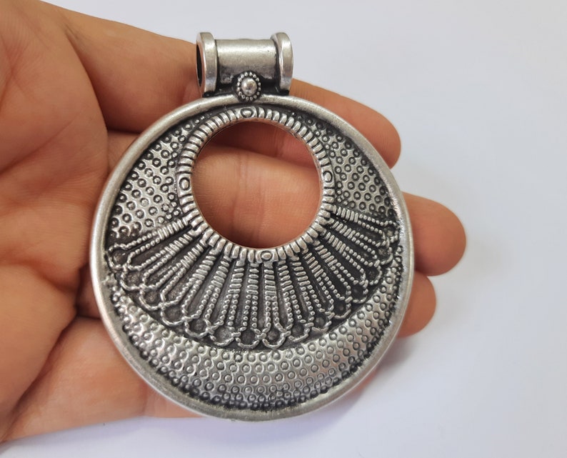 May include: Silver pendant with an intricate, circular design featuring a large open circle in the center. The pendant has a textured surface and a loop at the top for attaching a chain.