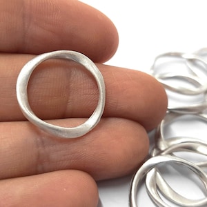 May include: A close-up of a silver ring with a unique, slightly wavy, organic shape. The ring is shown in a hand, with other similar rings in the background.