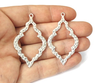 Puede incluir: Dos colgantes calados de color plateado con un acabado texturizado martillado. Cada colgante tiene forma de lágrima con un lazo decorativo superior para colgar. Los colgantes se sostienen en una mano sobre un fondo blanco.