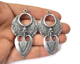 May include: A pair of silver-toned metal earrings with a circular design and a heart-shaped pendant. The earrings have a textured surface and a detailed design.