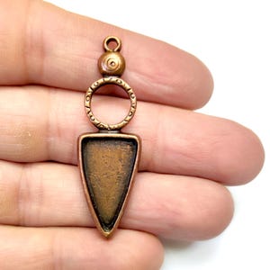 May include: A copper-toned pendant with a triangular frame and a circular ring at the top. The pendant has a textured surface and a slightly aged look.