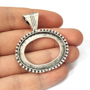 May include: An oval-shaped, silver-toned pendant setting with a textured border of small, raised dots. The pendant has a loop at the top for a chain and is designed to hold a stone or other insert. The setting is held in a hand.