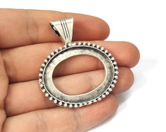 May include: An oval-shaped, silver-toned pendant setting with a textured border of small, raised dots. The pendant has a loop at the top for a chain and is designed to hold a stone or other insert. The setting is held in a hand.