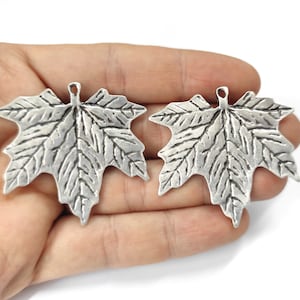 Peut inclure: Deux breloques en forme de feuille d'érable argentées avec une surface texturée. Les feuilles sont détaillées et ont un aspect réaliste.