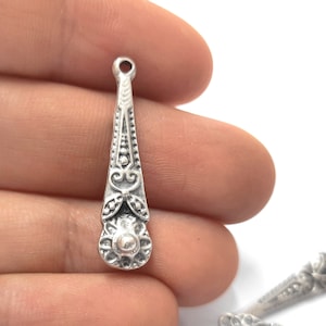 Puede incluir: Pendiente de plata con un diseño ornamentado. El pendiente tiene forma de lágrima y tiene una pequeña perla en la parte superior. El pendiente está decorado con intrincados patrones florales.