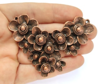 Könnte beinhalten: Ein kupferfarbener Metallanhänger in Form eines Blumenbüschels mit strukturierter Oberfläche. Der Anhänger besteht aus mehreren einzelnen Blüten, jede mit einer erhabenen Mitte und einem leicht gekräuselten Blütenblattdesign.