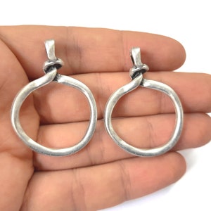 Puede incluir: Dos colgantes de metal plateado. Cada colgante presenta un diseño circular con un detalle de nudo en la parte superior. Los colgantes se sostienen en la mano de una persona sobre un fondo blanco. Los colgantes son probablemente para la fabricación de joyas.
