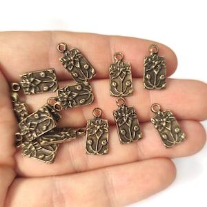 May include: A collection of 10 antique gold-toned metal charms featuring a floral design. Each charm is rectangular with a loop at the top for attaching to jewelry or other crafts.