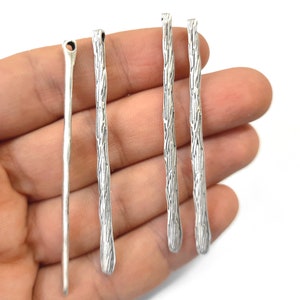 May include: Close-up of silver metal jewellery findings. The findings are long, thin, and have a textured, bark-like appearance. One finding has a hole at the top. The findings are held in a hand.