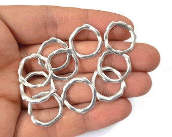 May include: A collection of silver-colored, irregular-shaped ring charms. The charms have a textured surface and a circular design with an uneven, wavy edge. The rings are held in a hand against a white background.
