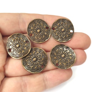 以下が含まれることがあります： 複雑な花柄が施された、アンティークゴールド調の金属製の丸い装飾チャームが5つ。各チャームには、プロジェクトに取り付けるための穴が2つあります。