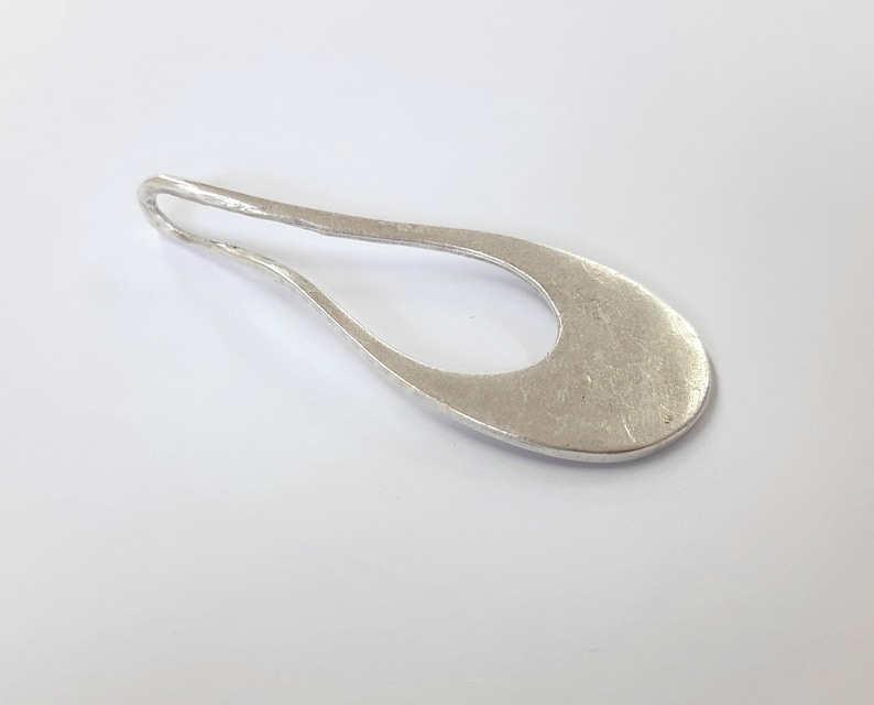 May include: A silver pendant with a smooth, oval shape and a hole in the center. The pendant has a textured surface.