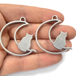 以下が含まれることがあります： 三日月型の猫のチャームが2つ。猫は三日月の縁に座っており、後ろ向きで、しっぽは三日月を巻いています。