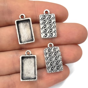 May include: Four silver-toned metal pendant charms. Two charms feature a rectangular frame with a smooth surface. Two charms feature a rectangular frame with a decorative swirl pattern.