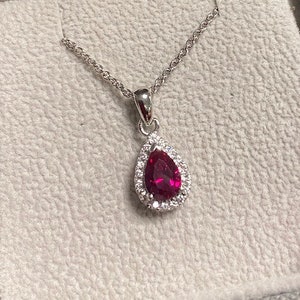 Könnte beinhalten: Eine silberne Halskette mit einem tropfenförmigen Anhänger, der einen roten Edelstein mit einem Heiligenschein aus kleineren weißen Edelsteinen zeigt.