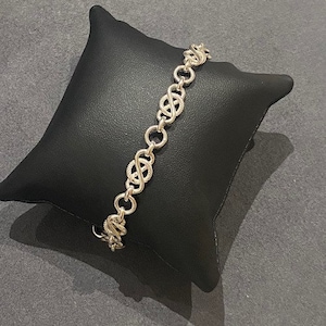 Könnte beinhalten: Ein silbernes Kettenarmband mit einem zarten, filigranen Design. Die Kette besteht aus kleinen, miteinander verbundenen Kreisen, die ein sich wiederholendes Muster bilden.