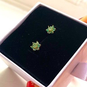 May include: A pair of gold earrings with green gemstone accents. The earrings are in a small white box.